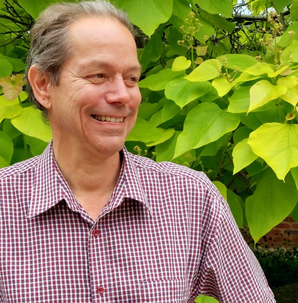 A portrait of Prof Simon Gilson FBA smiling infront of greenery. He is wearing a check shirt.