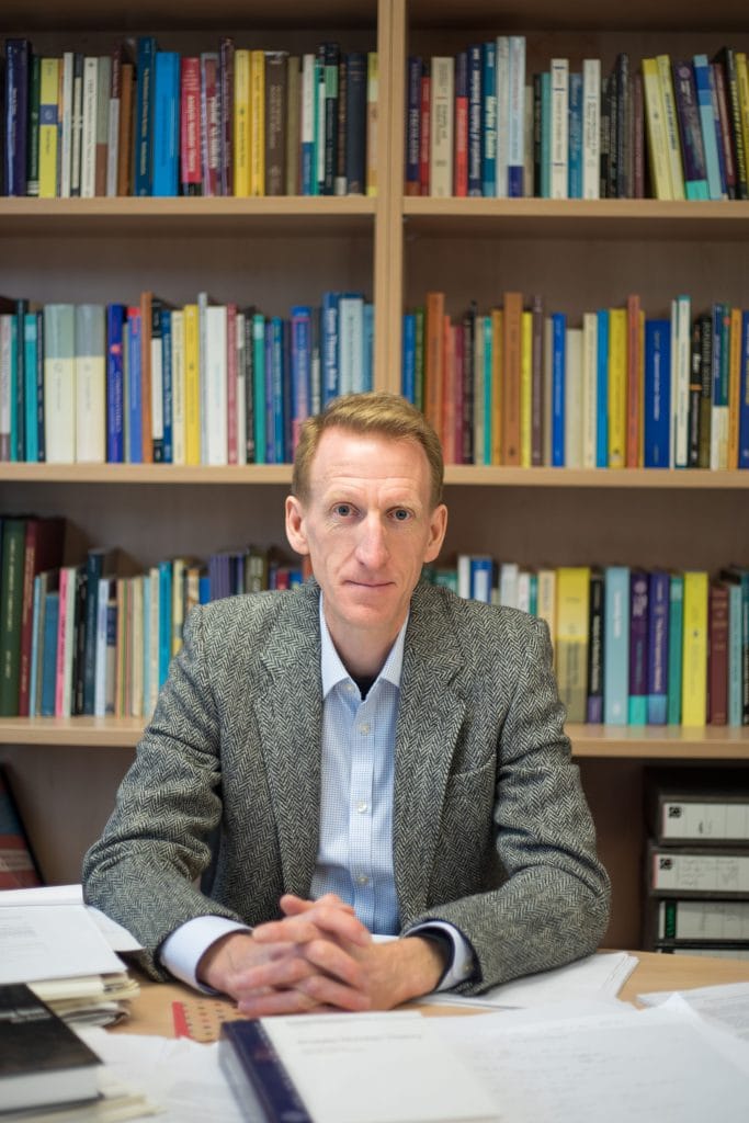 a oirtrait of ben green sitting at his desk with bookshelves behind him. he wears a tweed jacket and blue shirt and has his hands folded on the desk infront of him. He is smiling.
