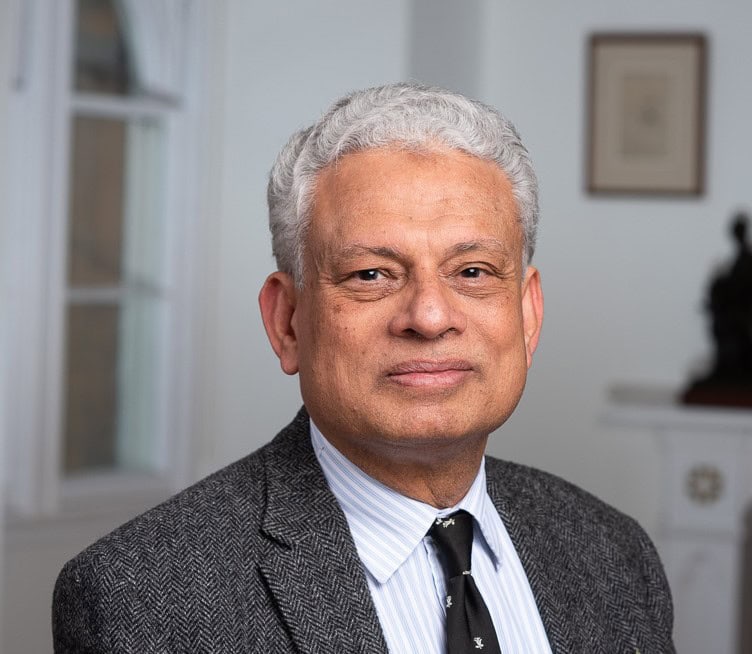 a portrait of Dr Farhan. He is wearing a grey tweed suit jacket, striped shrt, and magdalen tie. He is smiling.