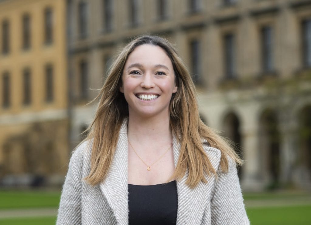 A portrait of Rachel wearing a tweed coat and a black top smiling in front of new buildings