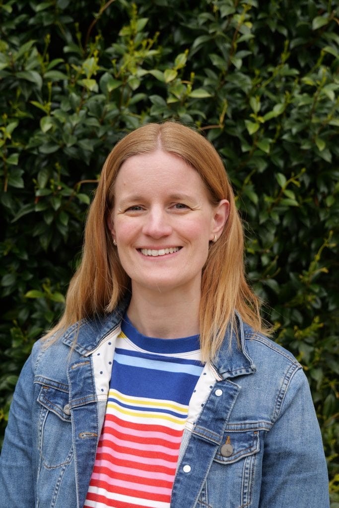 A portrait of Sarah smiling against a dark green leafy background