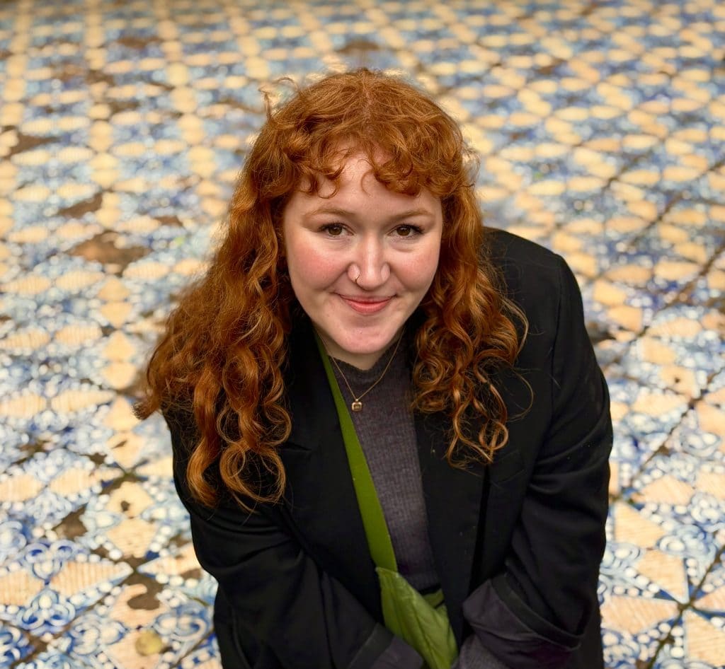A portrait of Lucy Wallis wearing a black jacket and a grey jumper. She has red curly hair and is smiling. There are tiles in the background