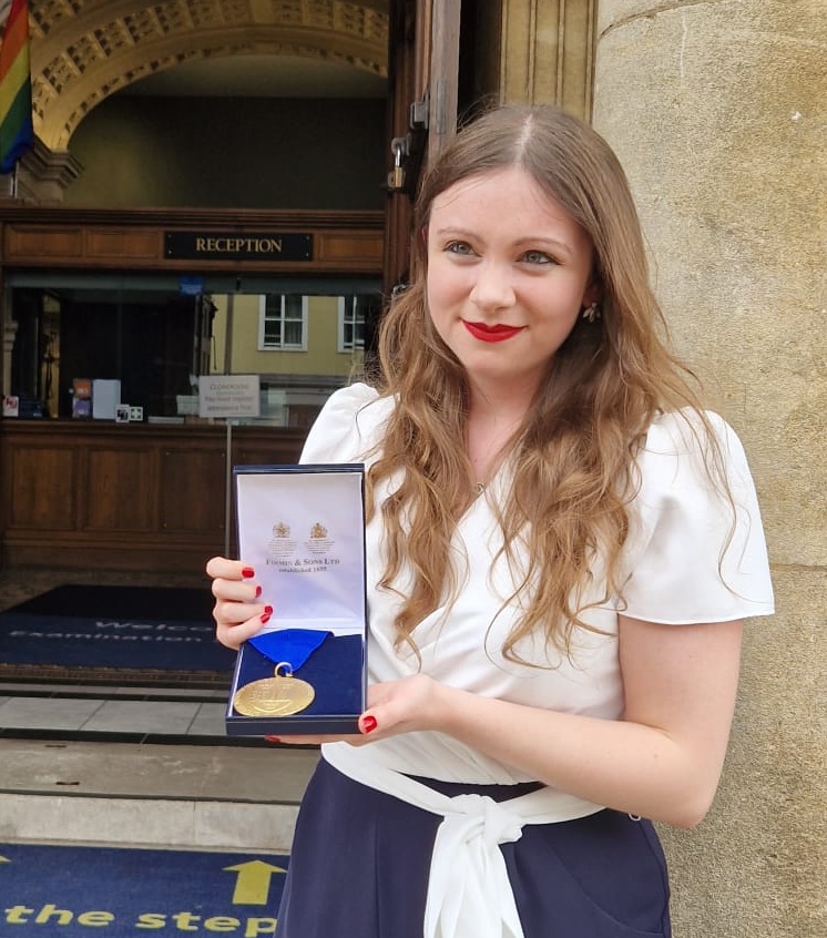 Scarlett is standing outside the Examination Schools on the High in Oxford, holding the Meakins-McClaran medal. It is a large gold disk on a blue ribbon, sitting on blue velvet, in a blue box.