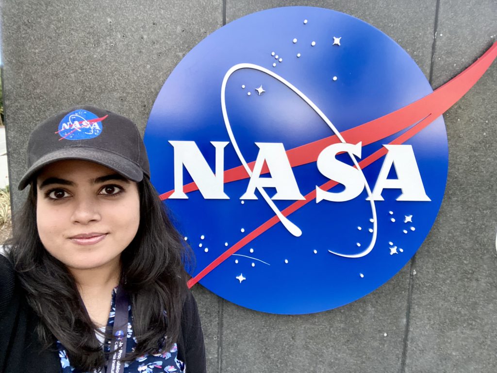 Hannah Rana stands next to the blue Nasa logo sign. She is wearing a black top and a black Nasa Cap.