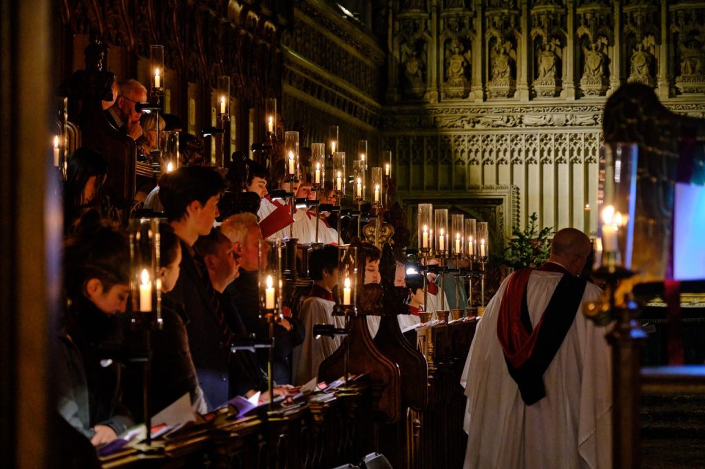 A photograph of the choir singing in chapel, they are bathed in warm candle light, and informator choristarum Mark Williams has his back to the camera while conducting.