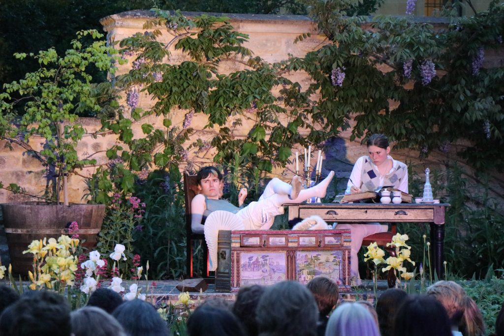 a photograph of Wally McCabe (Who played Orlando) and Anna McMullan (ensemble/ Sasha) on stage in the president's garden. Orlando lounges rouguishly in a chair, while Anna (as Woolf/Ensemble) writes in a large book at a writing desk. It is dusk, and the garden is suffused with a rosy glow The flowers are picked up by the light.