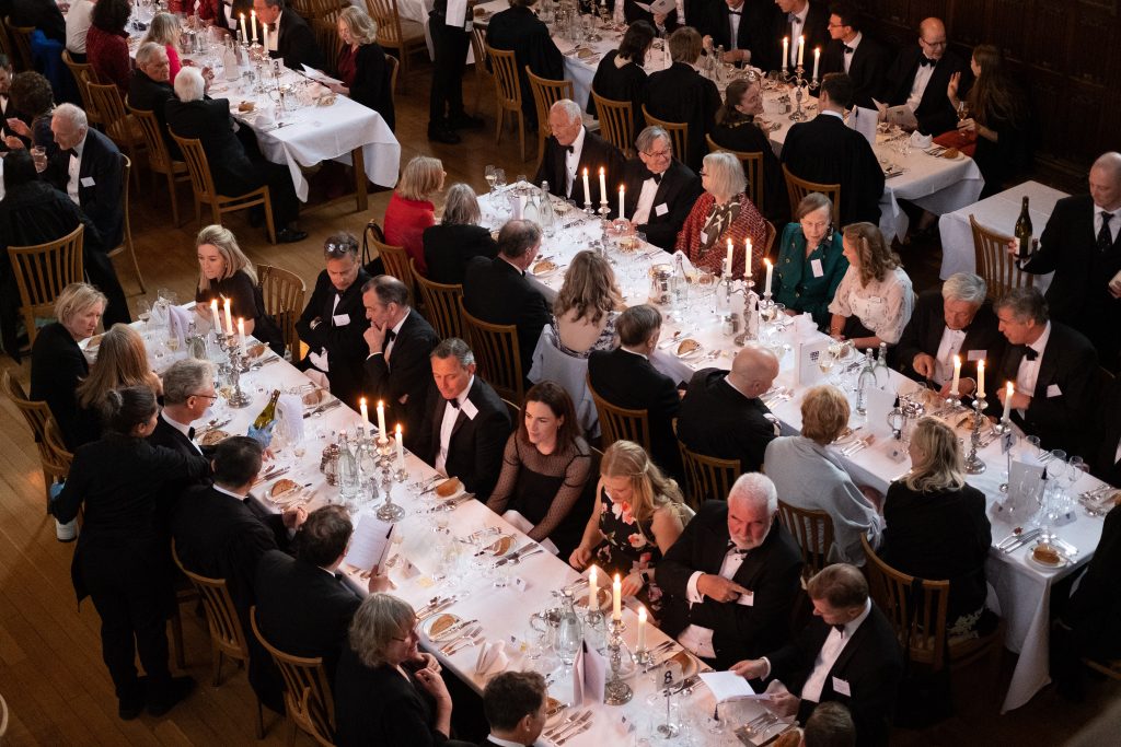 People sitting at white clothed tables that are fully laid. Everyone is dressed in Black Tie - on each table are lit candelabras.