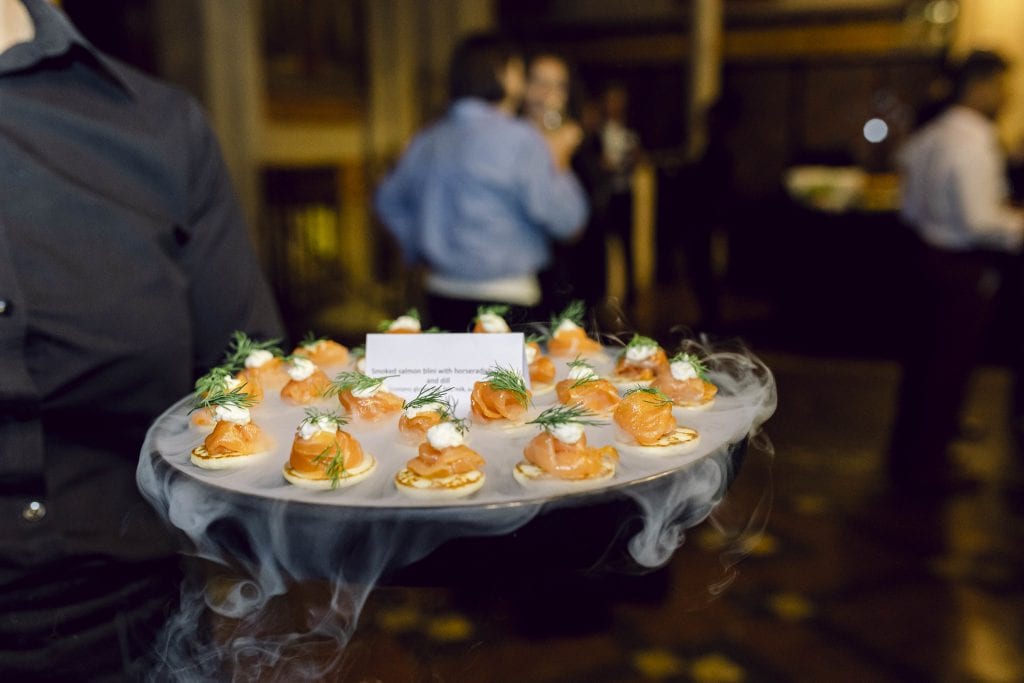 Salmon blini with horseradish and dill on a tray carried by a server - the tray is covered in smoking dry ice