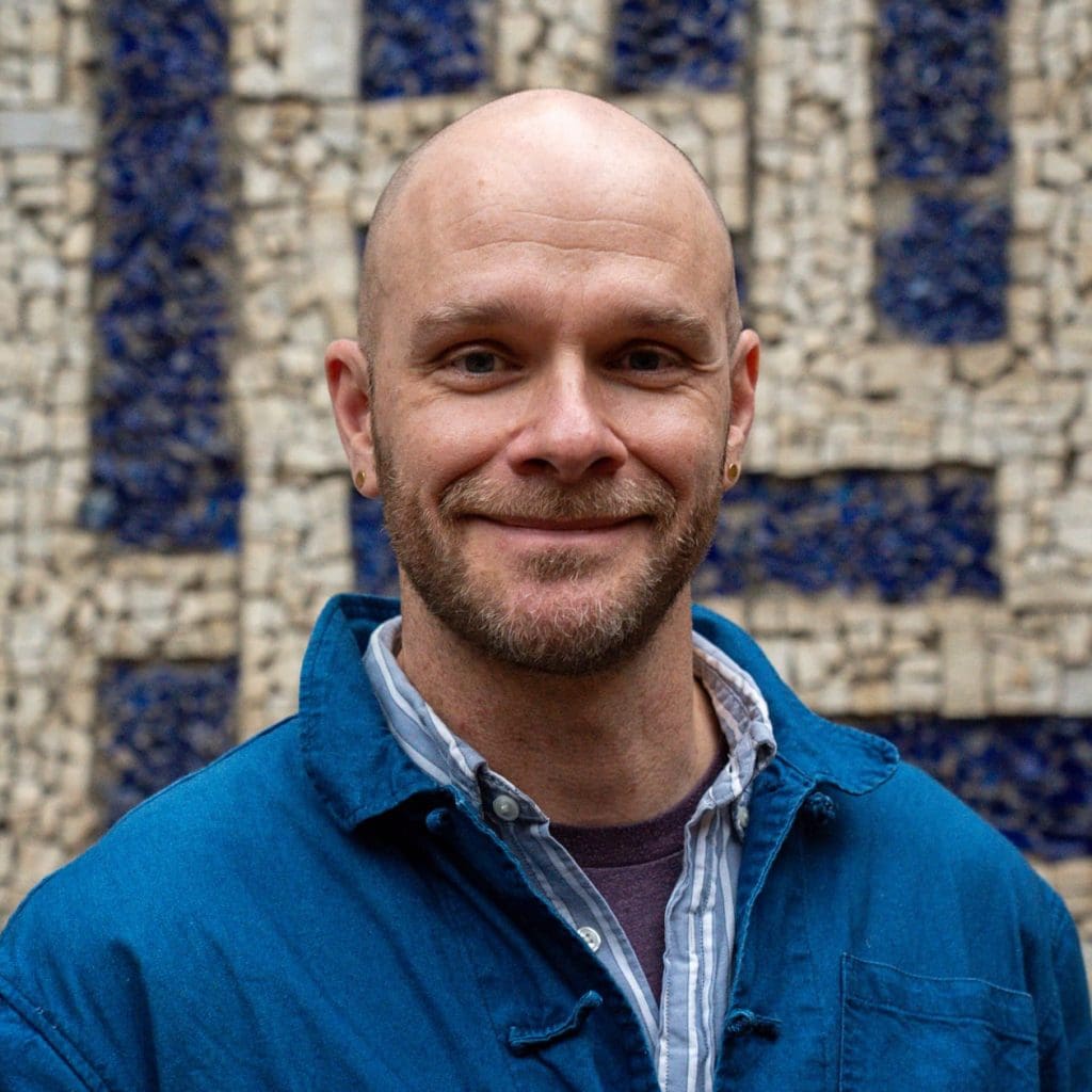 a portrait of pascal in front of a mosaic. He is smiling, has a beard, earrings, and is wearing a denim jacket and a blue striped shirt.