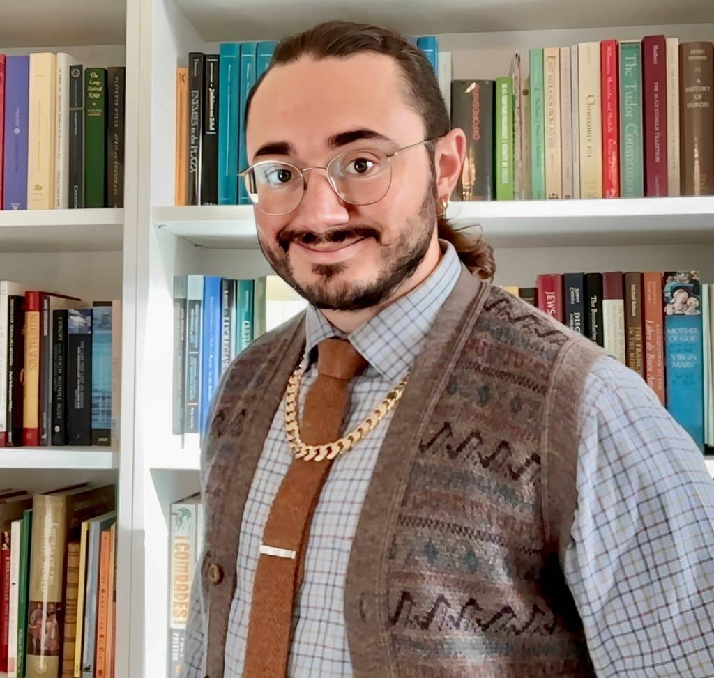 A portrait of Dr George Manning wearing a shirt, tie, and knitted vesy. He has long hair in a ponytail and is smiling in front of his bookshelves