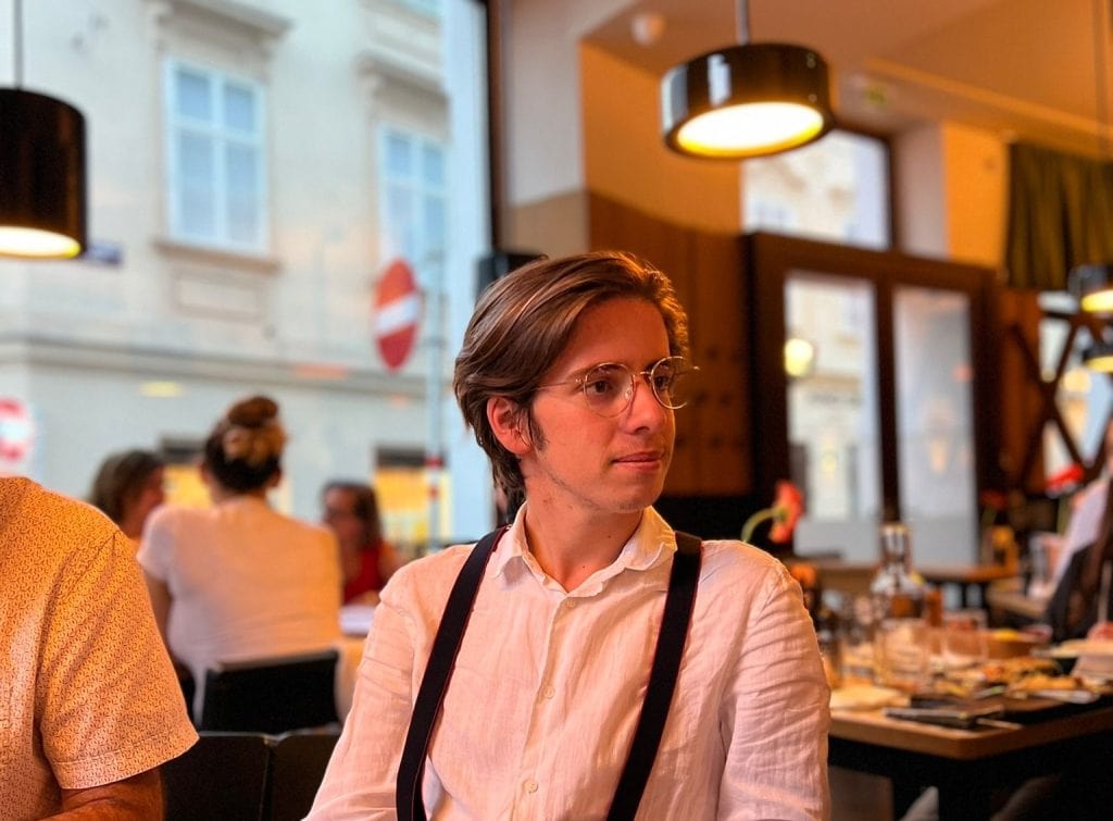 A portraite of antoine wearing a white shirt and braces, he has short brown hair and glasses.