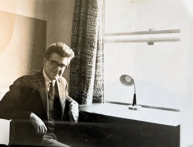A scan of a black and white photograph of Glyn in his bedroom in the Waynflete. He is seated at a desk and wearing glasses. You can see his lamp on the desk and some patterned curtains at the window. 