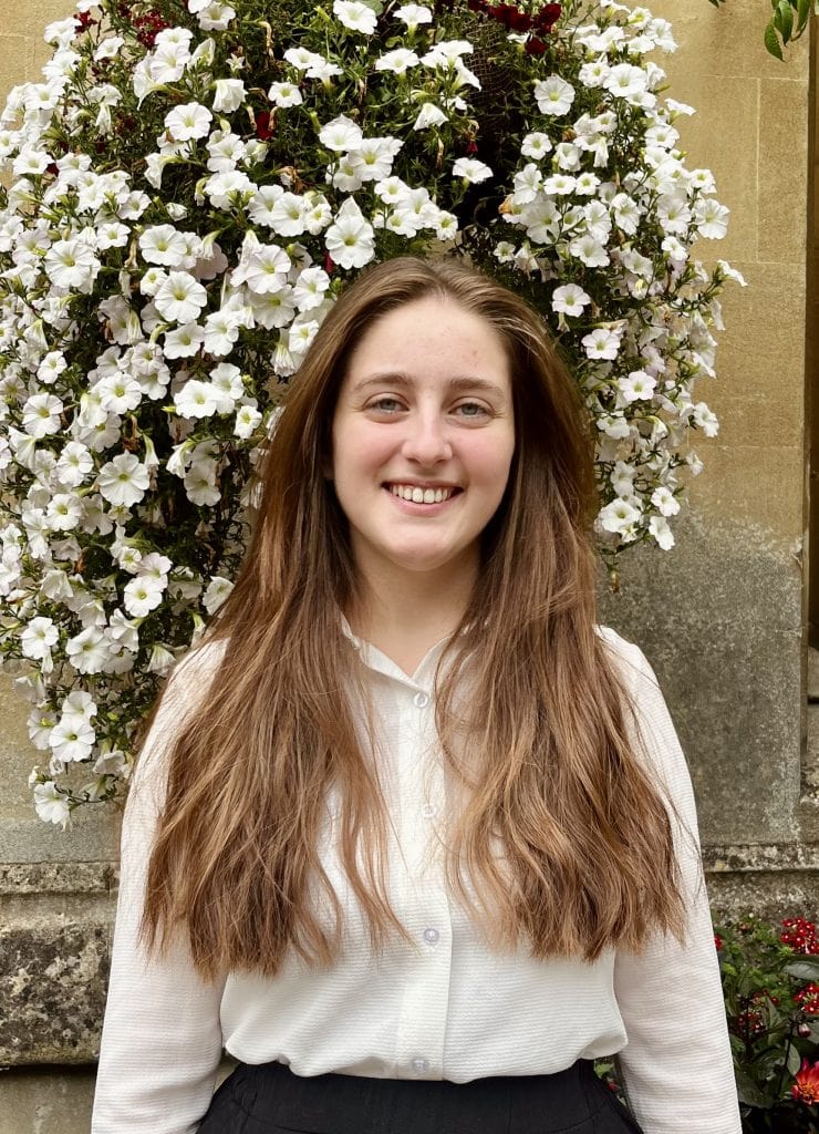 Portrait of Sofia standing in front of a hanging basket. She is wearing a white shirt and has long brown hair