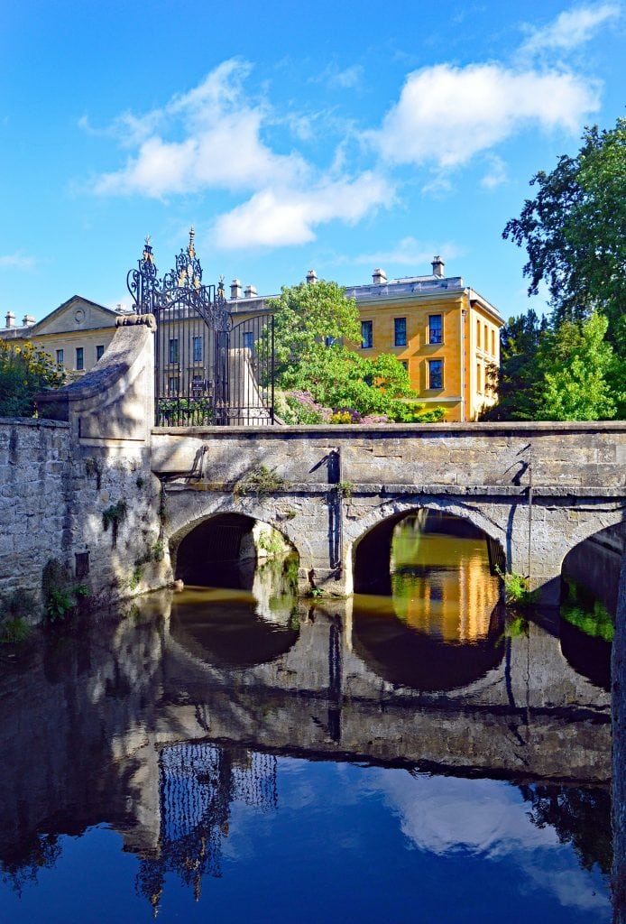 the view of New Buildings across the Cherwell in high summer. The bridge is reflecting in the rive, and the gate is open. In the distance you can see the verdent overflow of the herbaceous border.