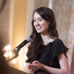 A portrait of Charis Jo speaking into a microphone. She has long dark hair and is smiling.