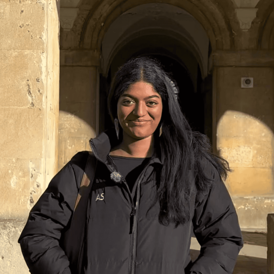 Aparna is wearing a Magdalen puffer, has long black hair, and is smiling at the camera. She has a warm smile, and the sunlight highlights her face. Shadows from the arches create an interesting contrast on the walls behind her.