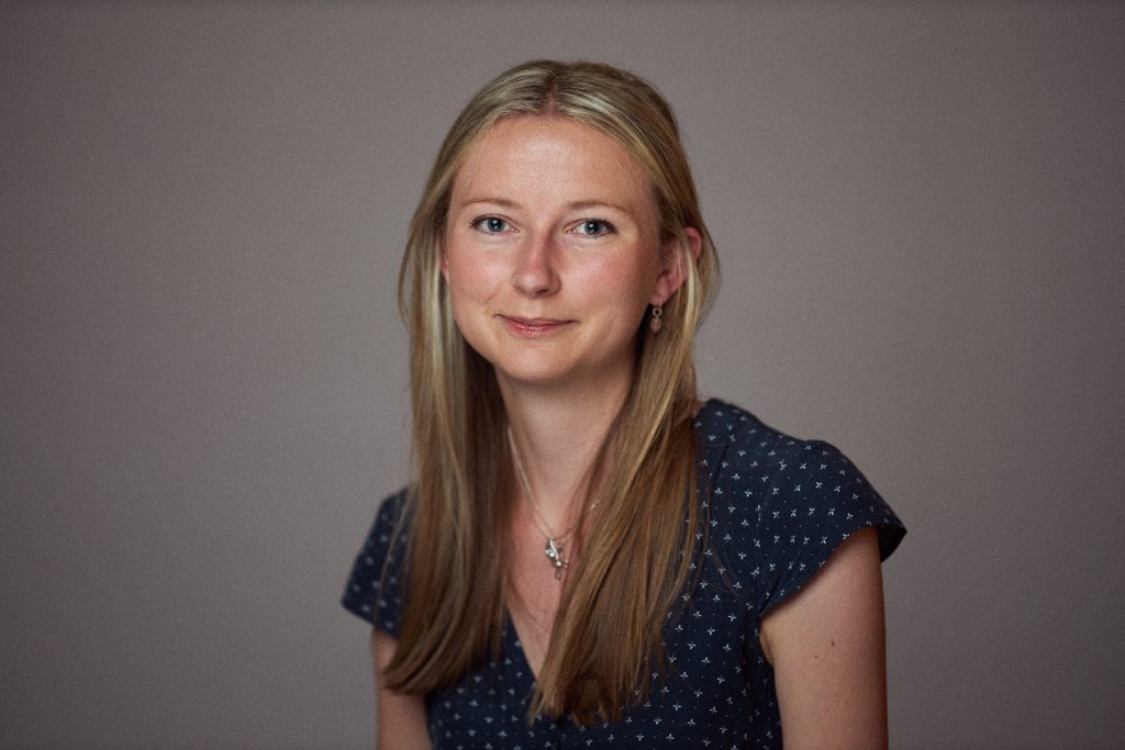 A portrait of rachel smiling at the camera. She has long blonde hair and wears a navy dress. The background is a neutral gray, and the lighting is soft and even, creating a natural and warm tone.