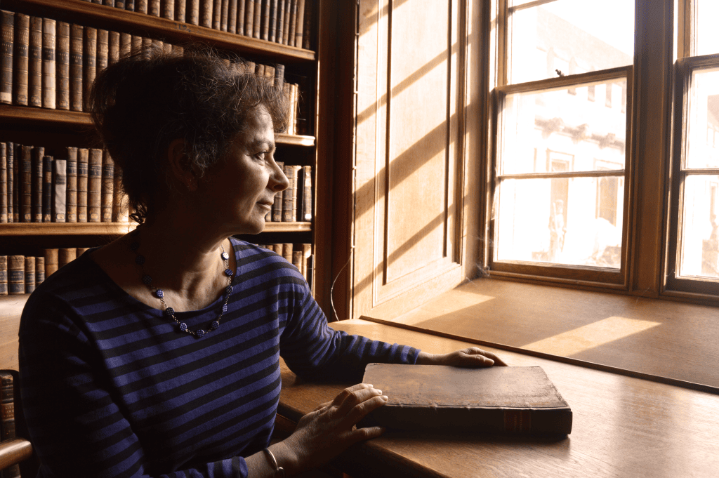 Anne Duprat sits at a wooden table in a historic library, gazing thoughtfully out of a sunlit window. She rests one hand on a large closed book in front of her. Shelves of antique leather-bound books fill the background, and warm natural light casts shadows across the wooden interior.