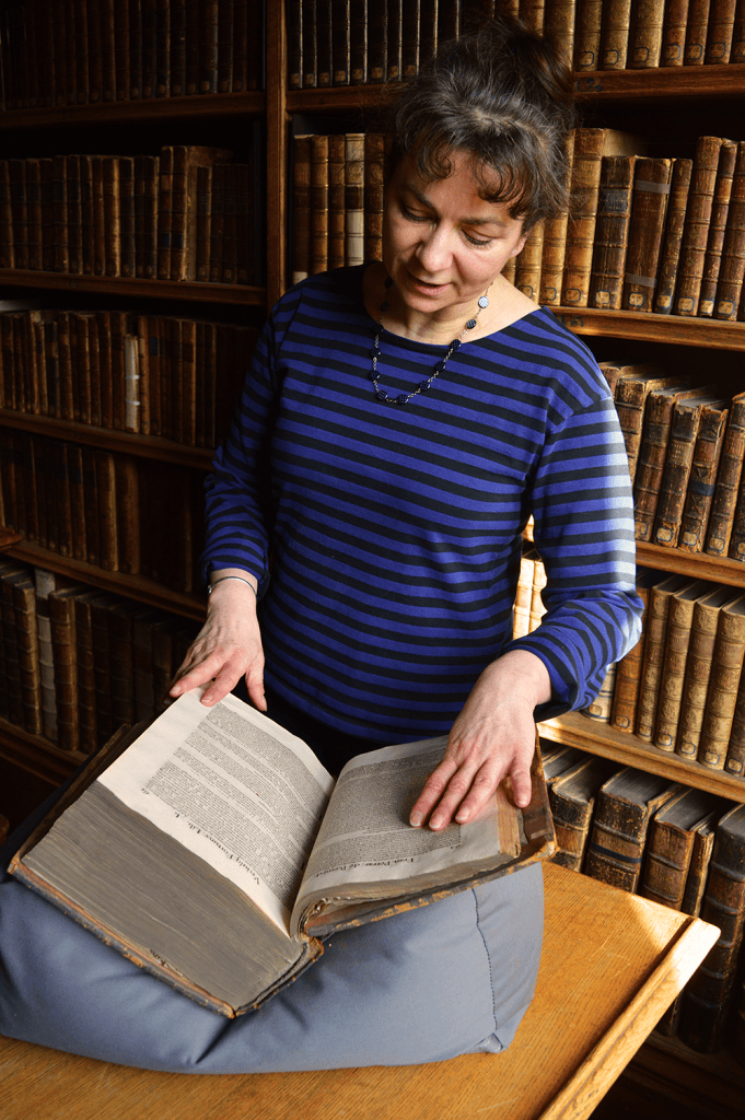 Anne Duprat stands in a historic library, looking down at a large open book resting on a cushioned support atop a wooden table. She is wearing a navy and black striped shirt and a beaded necklace. Shelves filled with old leather-bound books line the background, creating a scholarly, archival atmosphere.