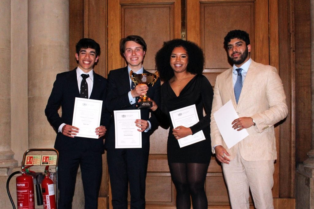 Four students from the University of Oxford standing in formal attire, holding certificates and a trophy after winning the 18th International Roman Law Moot. They are smiling and posing in front of wooden doors inside a historic building