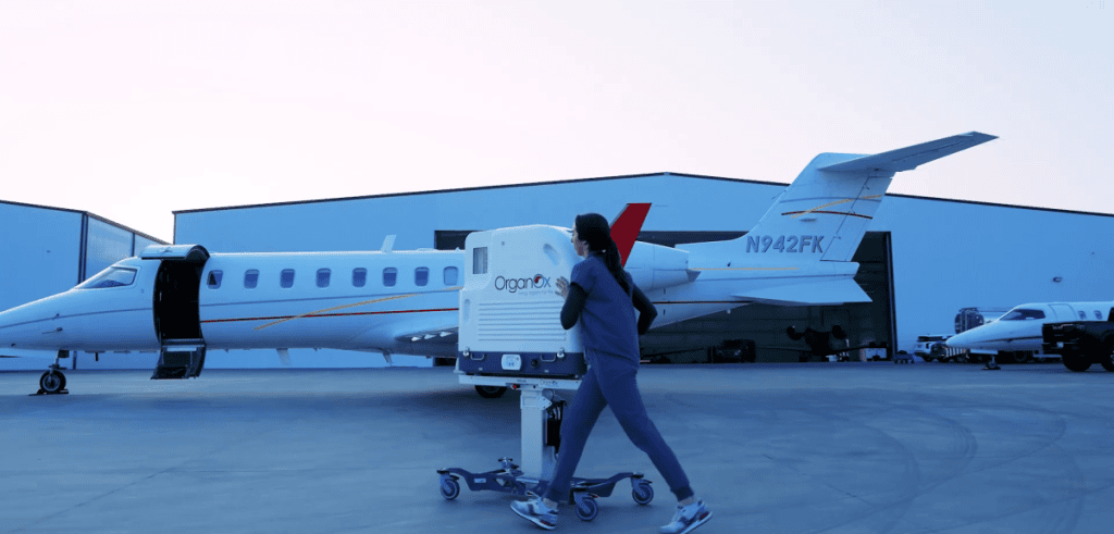 A medical professional in scrubs pushes an OrganOx normothermic perfusion device across an airport tarmac toward a private jet, illustrating the portability of the organ preservation technology for rapid, global transplant logistics.
