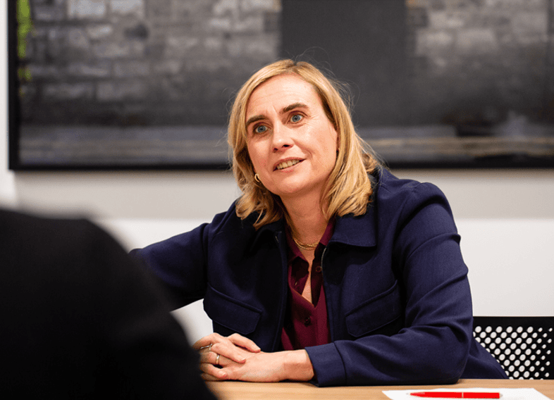 Sarah Healey with shoulder-length blonde hair wearing a navy blazer and burgundy blouse sits at a table, leaning slightly forward and smiling attentively during a conversation. A blurred figure is in the foreground, suggesting a meeting or interview setting.