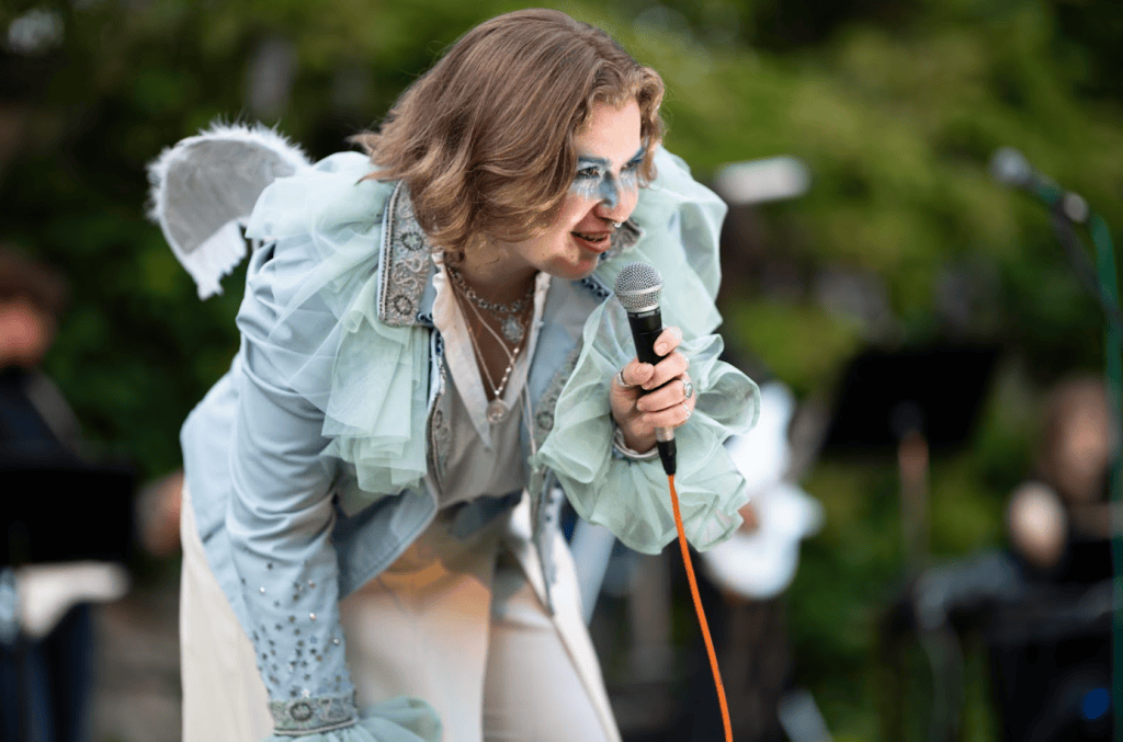 EP holding a microphone dressed in gauzy turqiose singing in the Tempest as Ariel