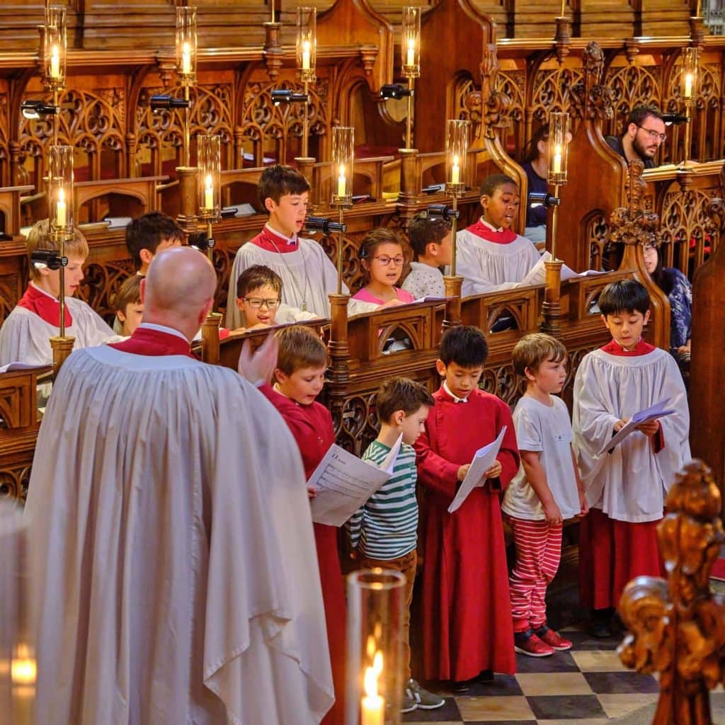 The Choir of Magdalen College, including potential new choristers, rehearses in an ornate wooden chapel, lit by glowing candle-like lamps. The choristers, some in red cassocks and others in white surplices, stand in carved wooden stalls, singing and holding sheet music. A conductor in a white robe leads from the front, gesturing with his hand. The setting is rich with gothic architecture and warm lighting, creating a reverent and focused atmosphere.