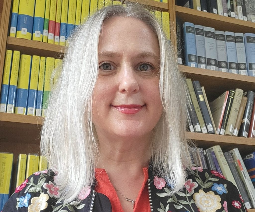 Claire Robe with white hair and a floral jacket standing in front of bookshelves.