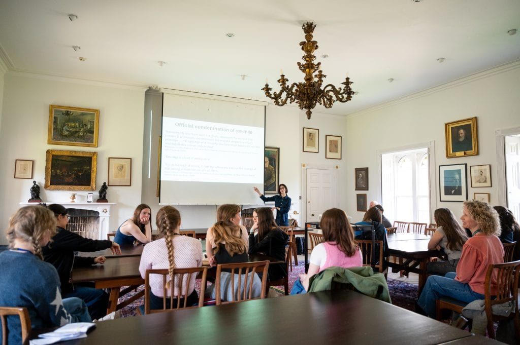 A group of students sit looking at a woman pointing to a projected PowerPoint slide.