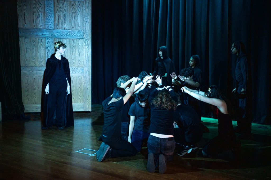 A boy in crown and black cloak looks dramatically at three hooded figures stood behind a circle of young actors kneeling with their hands outstretched into the middle.