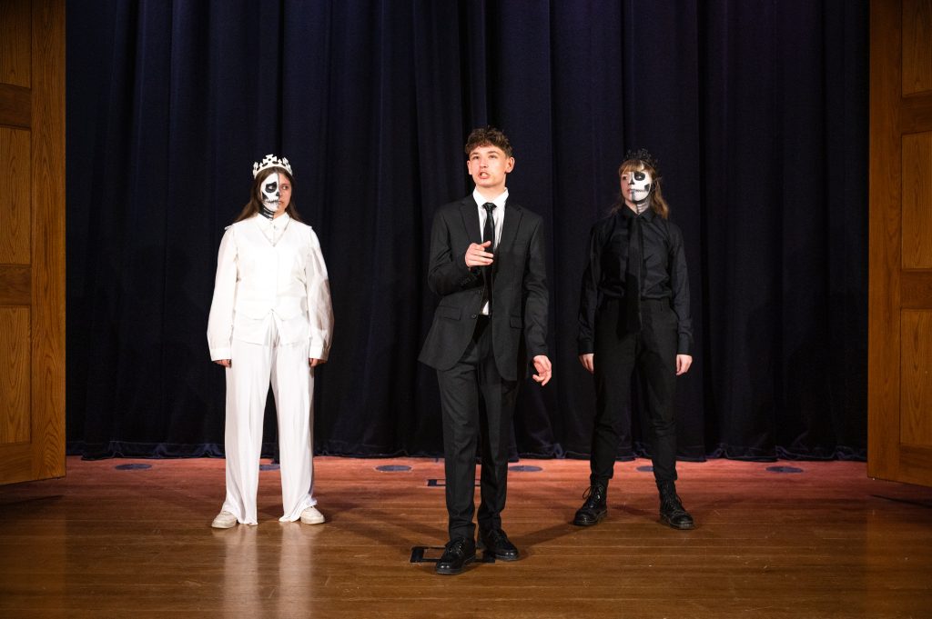 A boy stands centre stage mid-speech. Two girls stand either side looking at him, one dressed in white and one in black, both with half their face painted as a skull.