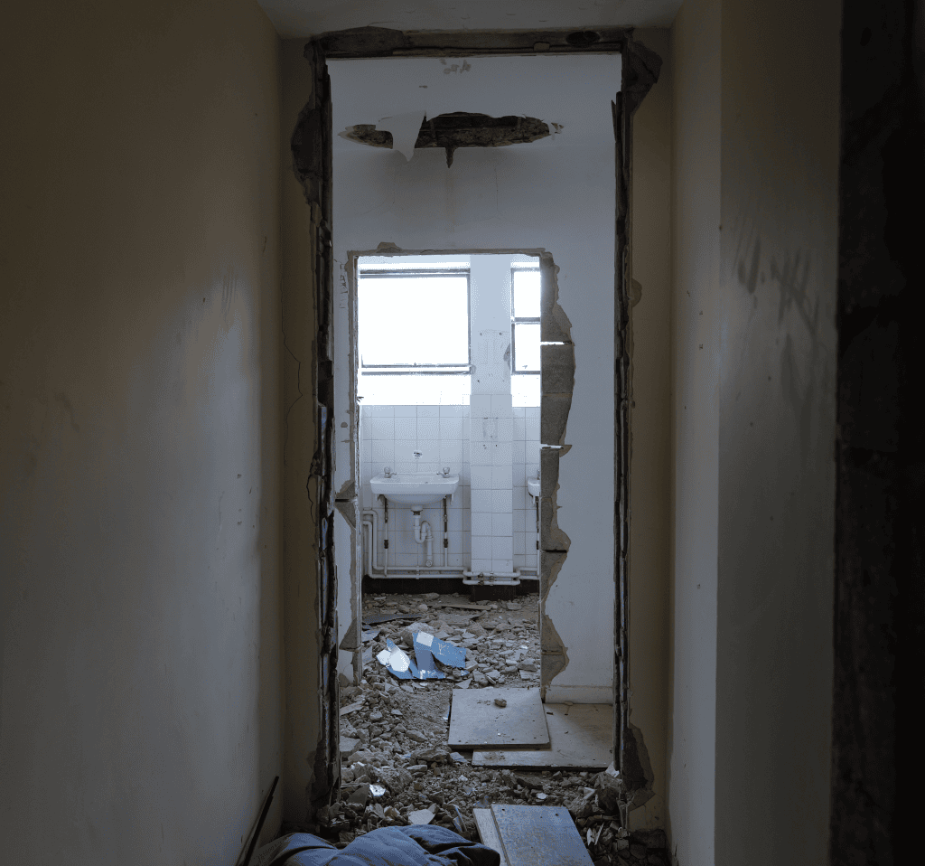 A narrow, dimly lit corridor in the Waynflete building leads to a partially demolished bathroom. The doorway’s plaster and bricks are broken away, exposing rough edges and debris scattered across the floor. Light from two small windows in the tiled bathroom illuminates the scene.