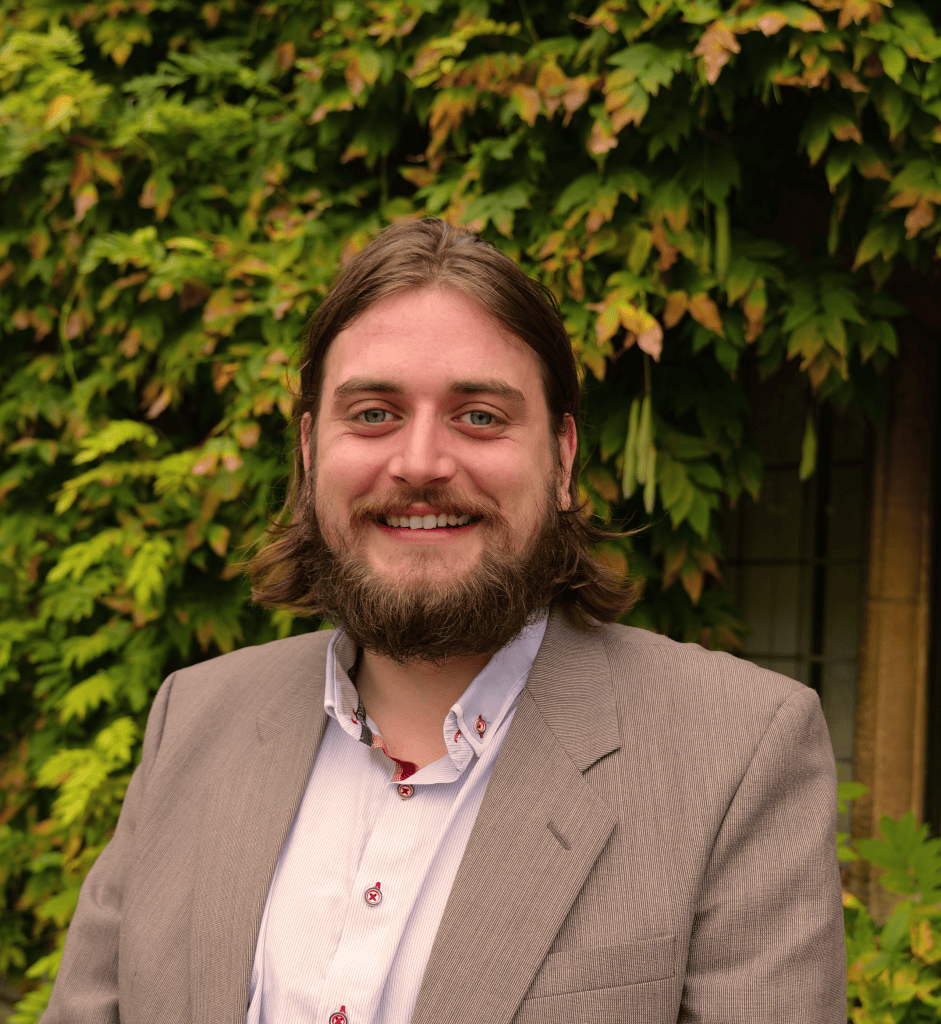 a PORTRAIT OF callum hunt smiling against a backdrop of green leaves. He is wearing a suit jacket, a shirt with an open collar, and smiling warmly at the camera