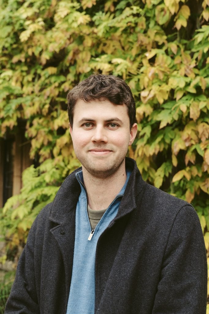 A portrait of Toby standing in front of autumnal leaves smiling wearing a blue jumper