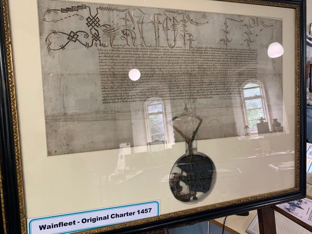 Framed image of the Magdalen Charter: The document features dense, handwritten Latin script in dark ink across the top half. Hanging from the bottom of the document by a green cord is a heavy, dark circular wax seal enclosed in a protective circular cutout in the matting. The document is framed in a dark wood frame with a wide, white matte border. Reflections of a window and indoor lighting are visible on the glass covering the document.