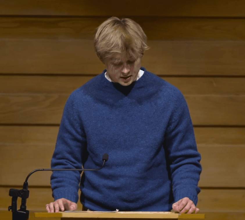 Angus Barrett, a young man with blonde hair, stands at a wooden lectern in front of a wood-paneled wall. He is wearing a textured blue knit sweater over a white collared shirt, looking down at his notes while speaking into a thin black microphone.