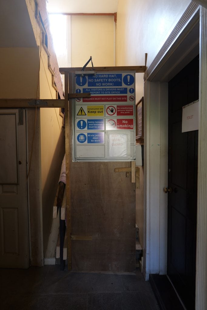 A temporary plywood door blocks a narrow hallway and staircase inside a building at Magdalen College, Oxford. A large blue and white safety board on the door lists site rules including "NO HARD HAT, NO SAFETY BOOTS, NO WORK!" and "Construction site Keep out." A black door to the right has a sign reading "TOILET."