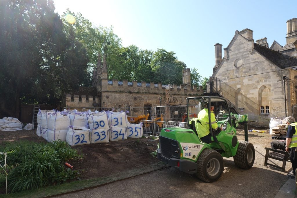 A green Weaver Plant loader operates in a courtyard at Magdalen College, Oxford. Large white industrial bags numbered 30 through 35 sit stacked on a dirt patch near stone buildings and mature trees. A worker in a high-visibility yellow jacket operates the vehicle. Text on the loader reads: "WEAVER PLANT HIRE & SPECIALIST GROUNDWORKS."