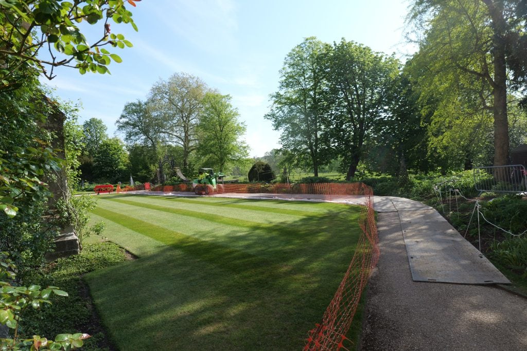 A manicured striped lawn at Magdalen College, Oxford, is protected by orange safety fencing and temporary ground protection mats. A small green loader is visible in the distance near an ornate iron gate. The scene is framed by lush green trees under a clear blue sky
