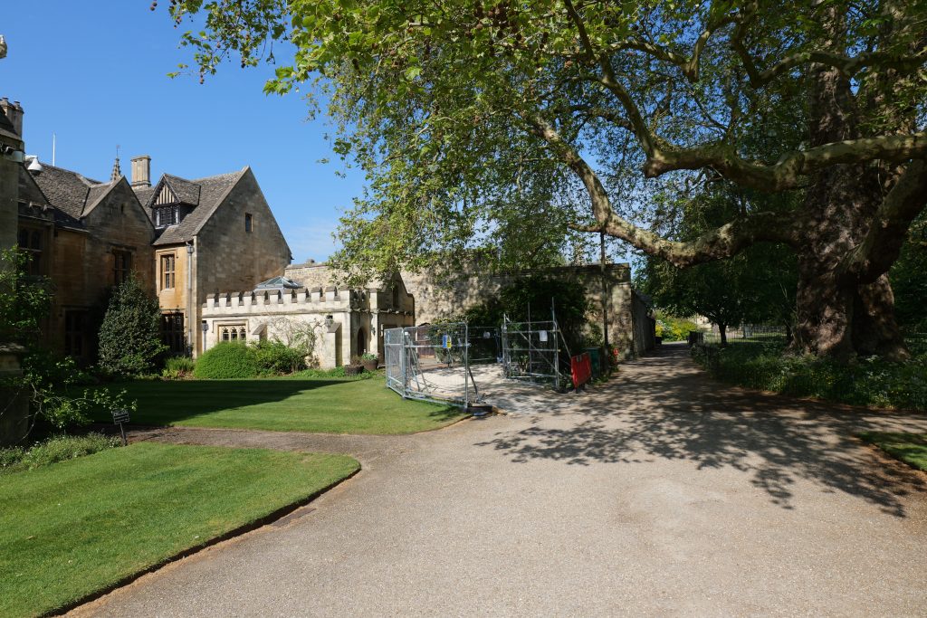 A gravel path at Magdalen College, Oxford, leads toward a small stone building with a crenelated roofline, partially enclosed by metal security fencing. To the right, a massive, ancient plane tree casts shade over the walkway. A small "PRIVATE" sign stands on a perfectly manicured lawn in the foreground. The sky is clear and bright blue.