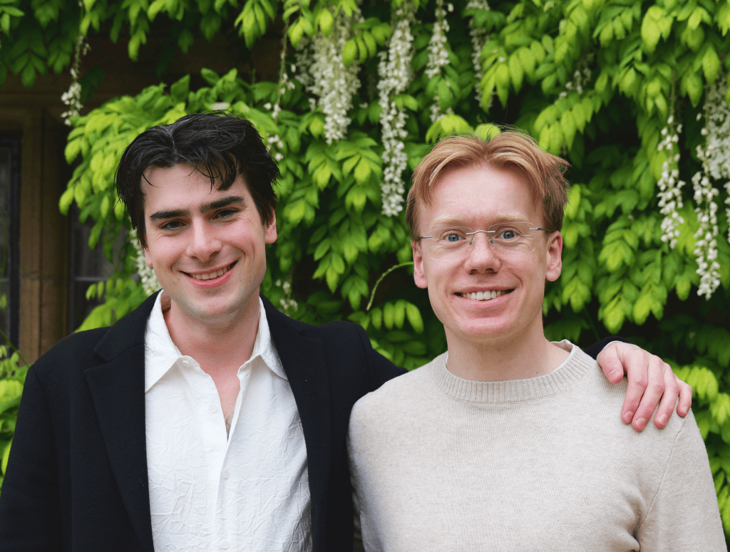 Two smiling young men stand side-by-side outdoors. The man on the left, Alex, has dark hair and wears a black jacket over a white collared shirt. The man on the right, Ollie, has fair reddish-blonde hair and glasses, wearing a cream sweater. An arm is draped over Ollie's shoulder, set against a dense backdrop of green leaves and white wisteria.