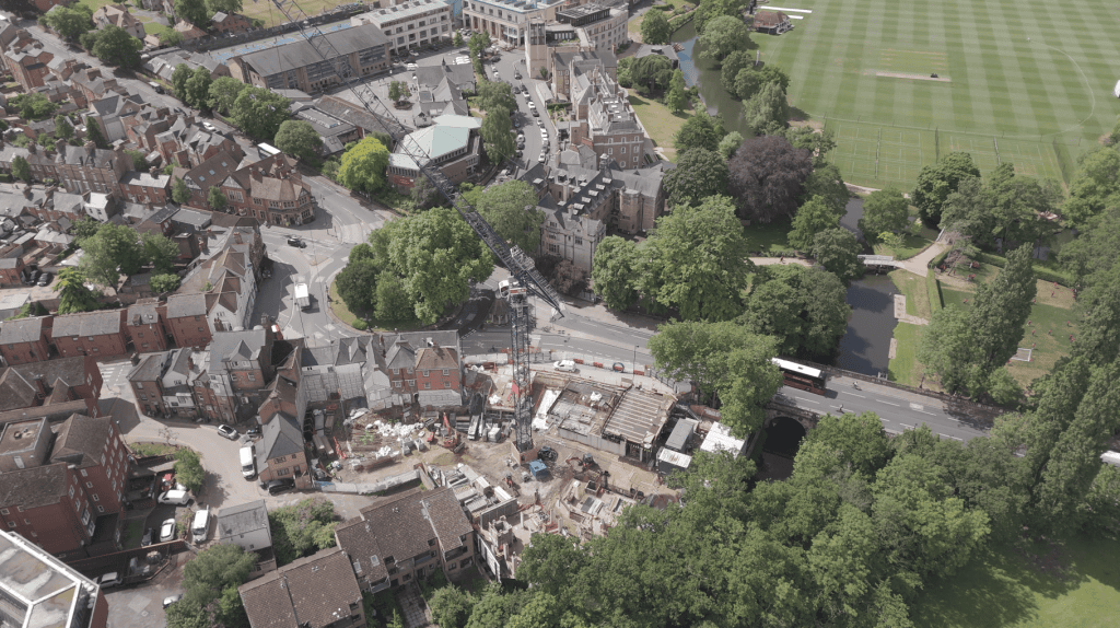 An aerial, bird’s-eye view captures the active construction site of the new Waynflete Quad development at Magdalen College, Oxford. A large blue tower crane stands at the center of the excavated foundations, surrounded by scaffolding, concrete footings, and construction materials. The site is bounded by St Clement's Street and the River Cherwell, which flows past Magdalen Bridge on the right. Lush green trees and the historic cityscape of Oxford encircle the development area.
