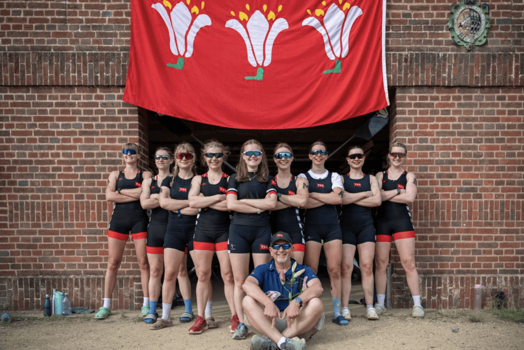Nine female rowers from Magdalen W1 rowing team stand in front of a brick boathouse entry under a large red flag featuring three white lilies. They wear black rowing kit with red accents and sunglasses. A coach sits cross-legged on the ground in front of them holding a flower.