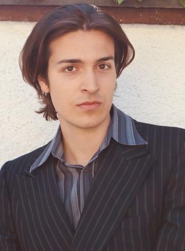 A portrait of Julian Kekulé. He has medium-length, dark brown hair parted down the middle and dark eyes. He is wearing a dark, pinstriped suit jacket over a grey and blue striped collared shirt. He poses against a plain, light-coloured textured wall.
