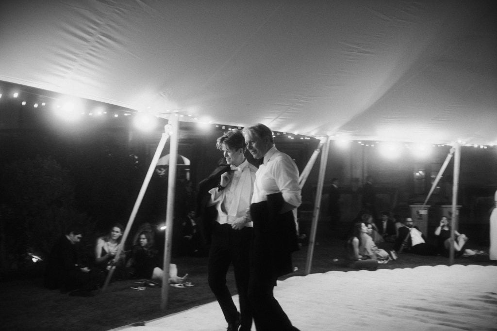 Black and white photograph capturing two men dressed in black-tie attire walking side-by-side across a light floor space. Warm fairy lights glow overhead along the interior of a large marquee tent. Guests sit around the edge of the lawn watching in the background.