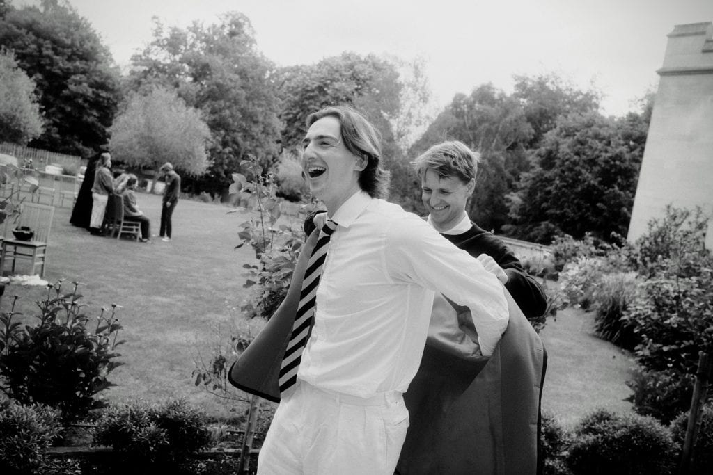 Black and white photo of a smiling young man in a white shirt and striped tie laughing as a friend helps him put on a blazer outdoors.
