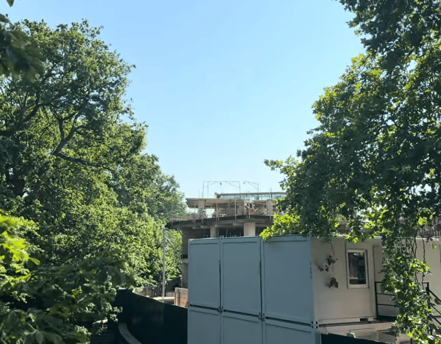 A building site peeking through a dense canopy of green summer trees. White modular site cabins sit in the lower right foreground. Behind them, a concrete building is under construction. It features scaffolding and exposed levels. The structure is partially visible between the large leafy branches. A clear, pale blue sky fills the upper portion of the frame.