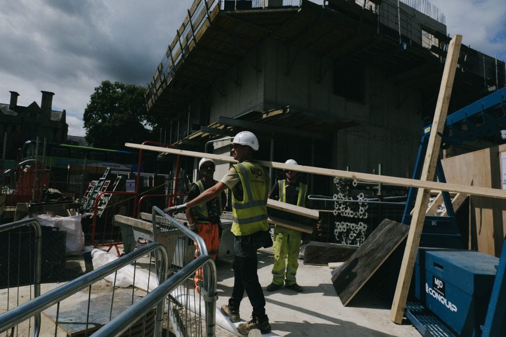 The busy Waynflete Quad construction site under a cloudy sky. Three workers wear hard hats and high-visibility vests. In the foreground, one worker walks across the concrete ground while balancing a long wooden beam over his shoulder, surrounded by metal barriers, scaffolding, and the concrete framework of a multi-storey building.