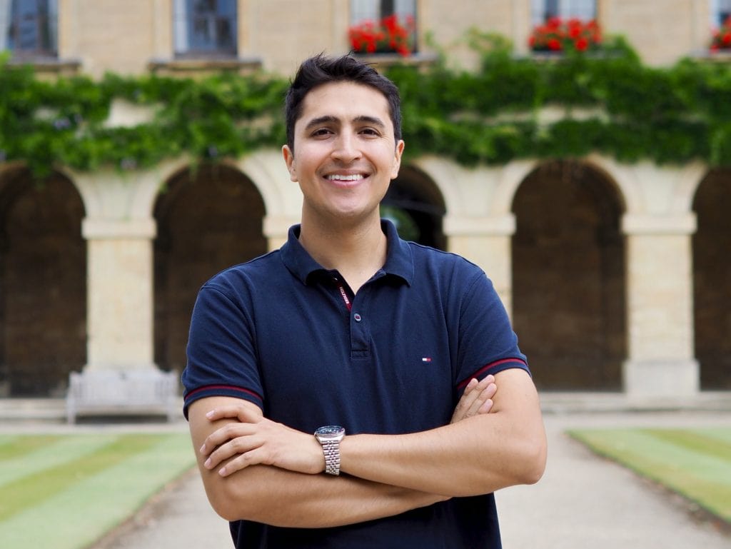 Kevin stands smiling with his arms crossed. He wears a navy polo shirt. The stone arches of Magdalen College's New Buildings frame the blurred background. Bright red flowers bloom along the building's windows.
